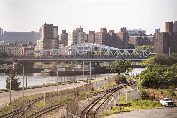 A silver bridge spans the Harlem River, with train tracks in the foreground and many brown and beige buildings on the other side of the river.