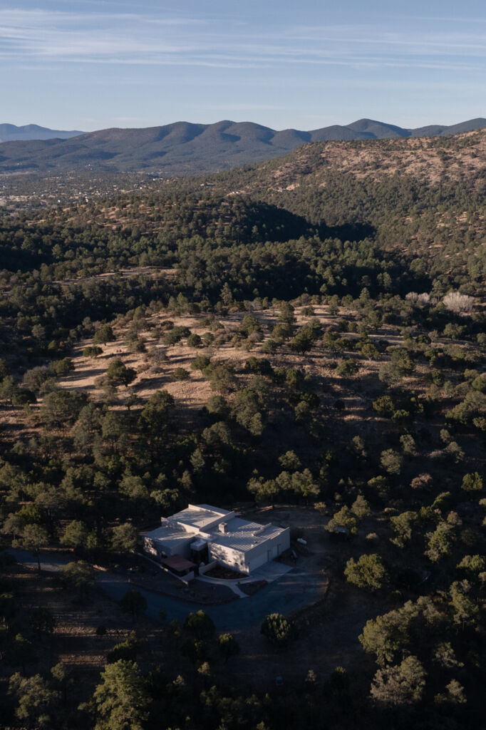 A large home is seen surrounded by a vast forest, with mountains in the background.