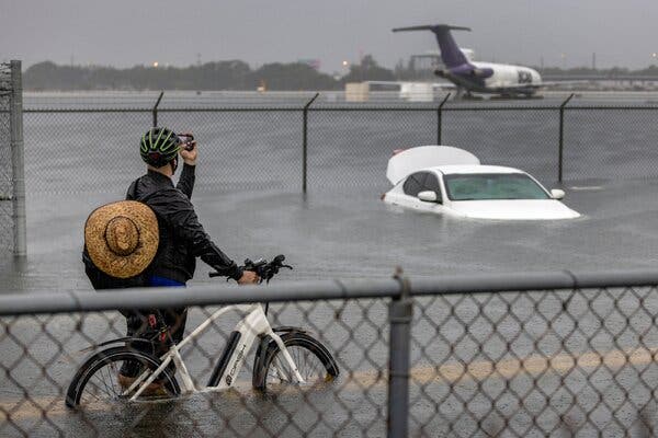 A person holding a bicycle stands and takes a picture of a plane on a runway, with a car partially submerged by flood water.